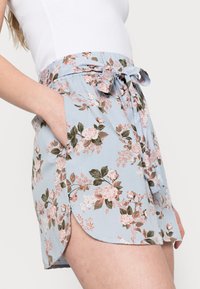 Kvinna klädd i ljusblå blommiga shorts med knytmidja och en vit ärmlös topp, med handen i fickan, mot en enfärgad bakgrund.