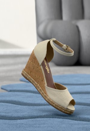 Sandale compensée beige avec bride à la cheville, talon en liège et semelle en jute tissé reposant sur un tapis texturé bleu à l'intérieur.
