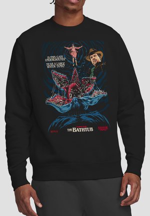Person trägt schwarzen Pullover mit einem roten Blumenmonster, zwei Charakteren und Text, der auf Stranger Things Kapitel Sieben: Die Badewanne verweist.