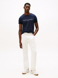 Marineblå bomulds-T-shirt med trykt logo, parret med hvide bukser og hvide sneakers med beige accenter. Modellen står med den ene hånd i lommen.