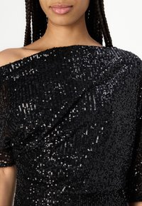 Top nero in paillettes con design off-shoulder, tessuto texturizzato e silhouette aderente. Caratterizzato da una finitura liscia con paillettes scintillanti su tutta la superficie.