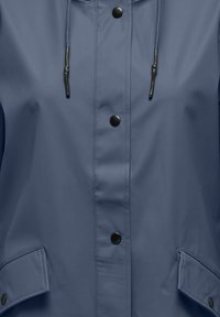 Modrá nepremokavá bunda s čiernymi cvokmi, nastaviteľnou kapucňou so sťahovacími šnúrkami a klopovými vreckami so zapínaním na cvoky.