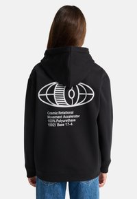 Persona che indossa un hoodie nero con un globo astratto bianco e testo che recita "Acceleratore di Movimento Rotazionale Cosmico 100% Poliuretano 1992/Base 17-4" sulla schiena.