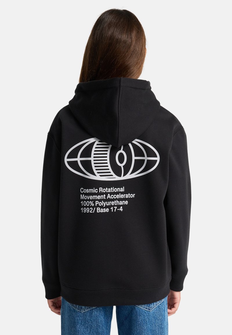 Persona che indossa un hoodie nero con un globo astratto bianco e testo che recita "Acceleratore di Movimento Rotazionale Cosmico 100% Poliuretano 1992/Base 17-4" sulla schiena.