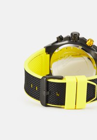Reloj cronógrafo negro con una correa de goma amarilla, que presenta un patrón texturizado, llamativos acentos amarillos y una parte trasera de acero inoxidable.