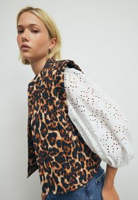 Luipaardprint vest met geplooid ontwerp, gecombineerd met een witte top met oogjespatronen en pofmouwen. Casual stijl met denim jeans.