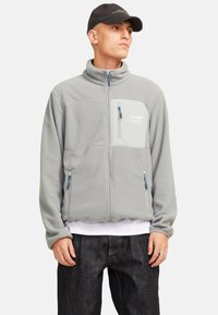 Chaqueta de forro polar gris con un cuello alto, cierre frontal y un bolsillo en el pecho en un tono más claro. Presenta cremalleras azules y detalles de costura.