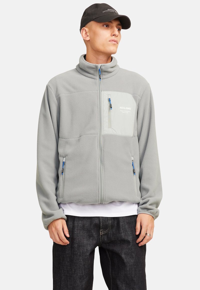 Chaqueta de forro polar gris con un cuello alto, cierre frontal y un bolsillo en el pecho en un tono más claro. Presenta cremalleras azules y detalles de costura.