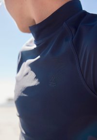 Tumesinise lühikeste varrukatega kaitsev särk kõrge krae, siledapoolse tekstuuri ja kergema tooniga minimalistliku logoga rinnal.