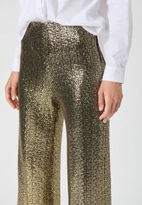 Pantalons en sequins dorés et noirs taille haute avec un motif texturé, une silhouette ajustée et des poches latérales, associés à une chemise blanche à boutons.