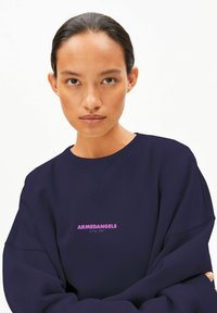 Marineblauer Pullover aus weichem Stoff, mit einem gerippten Kragen und einem rosa Logo auf der Vorderseite, auf dem "ARMEDANGELS since 2007" steht.