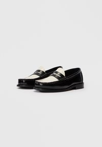 Um par de mocassins de couro preto e branco com saltos baixos e detalhes cosidos sobre um fundo claro.