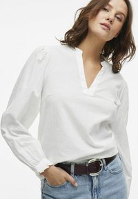 Hvid langærmet bluse med V-hals, tekstureret stof og rynkede ærmer. Kombineret med en blå denimnederdel og et brunt bælte med en sølvspænde.