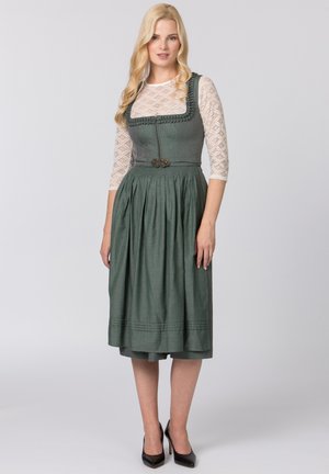 Grünes, knielanges Kleid mit taillierter Oberseite, gefälteltem Rock und dekorativem Besatz, kombiniert mit einem weißen Spitzenoberteil mit langen Ärmeln und schwarzen Absätzen.