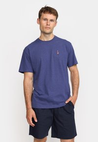 Blue heathered T-shirt in soft fabric with a crew neckline, featuring a small red and white graphic logo on the chest. Paired with dark shorts.