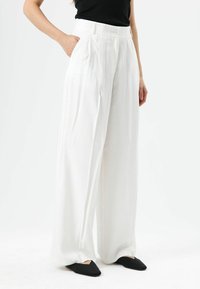 Pantalon blanc à jambes larges, taille haute, avec plis et poches latérales, fabriqué dans un tissu léger. Porté avec des chaussures noires sans lacets.