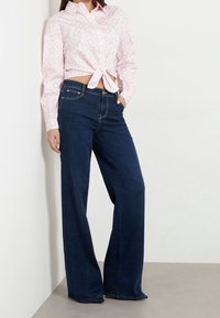 Vit och rosa mönstrad skjorta med knut i midjan, i kombination med mörkblå vida jeans. Slätt tyg och avslappnad passform.