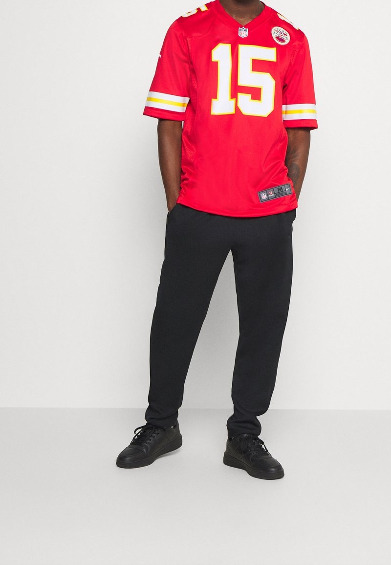 Homme portant un maillot rouge des Kansas City Chiefs NFL avec le numéro 15, pantalon noir et baskets noires, debout les mains dans les poches.