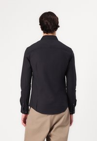 Camicia a maniche lunghe scura con un design aderente, caratterizzata da una spalla a vuoto, polsini con bottone e una texture del tessuto liscia.