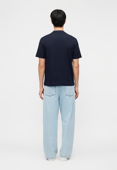 Άνδρας που στέκεται γυρισμένος ανάποδα, φοράει μια υφασμάτινη μπλε κοντομάνικη μπλούζα και ανοιχτόχρωμα τζιν με φαρδιά μπατζάκια και λευκά αθλητικά παπούτσια.