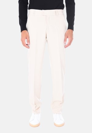 Uomo che indossa pantaloni su misura beige chiaro e sneakers bianche, in piedi contro uno sfondo semplice chiaro.