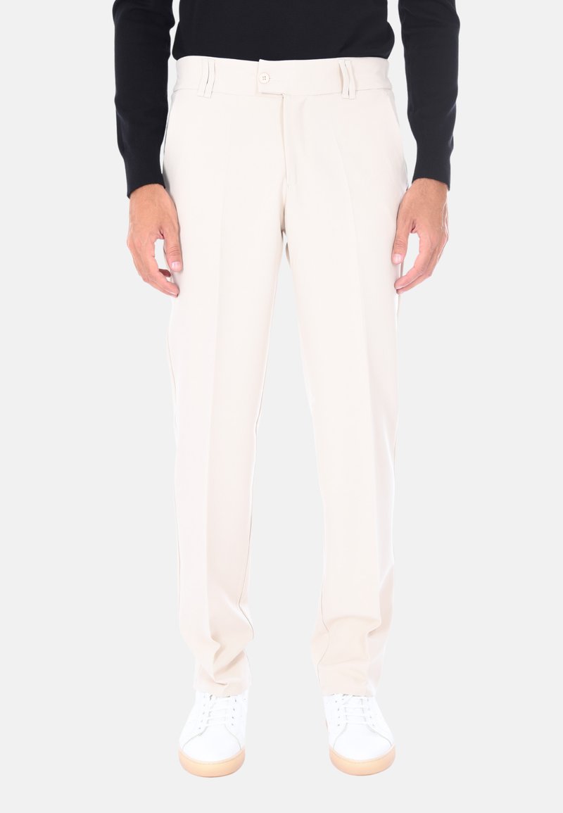 Uomo che indossa pantaloni su misura beige chiaro e sneakers bianche, in piedi contro uno sfondo semplice chiaro.