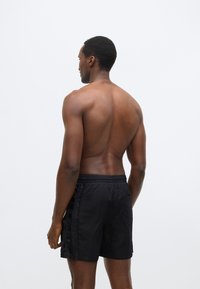 Μαύρο μαγιό σορτς από ελαφρύ ύφασμα, με ελαστική μέση και πλαϊνές τσέπες, διακοσμημένο με κάθετες λεπτομέρειες λογότυπου στο πλάι.