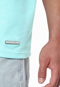 Lyseblå bomulls T-skjorte med korte ermer, med en sølv logo etikett i kanten. Grå shorts og en hånd synlig på siden.