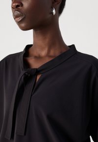 Zwarte blouse met een striknekdesign, textuurstof en een subtiele glans. Ronde halslijn met een sleutelgatopening aan de voorkant.