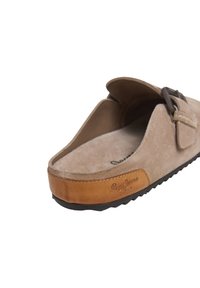 Bež semiš slip-on mule s kožnim potplatom, crnom potpunom i podesivim remenom na vrhu.