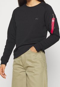 Sudadera negra hecha de una mezcla de algodón, con cuello redondo, mangas raglán y un bolsillo con cremallera roja en el brazo izquierdo.