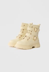 Botas de tornozelo bege feitas de material sintético, com formato curvado, fecho de atacadores na frente, duas tiras, detalhes em forma de coração dourado e sola texturizada.