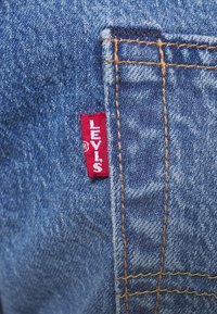 Blå denimtyg med en synlig röd etikett som säger "LEVI'S." Har gyllene sömmar och en fickdetalj på sidan.