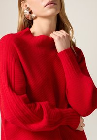 Maglione rosso a coste con collo alto e maniche lunghe con motivi testurizzati. L'accessorio include un orecchino circolare con accenti colorati.