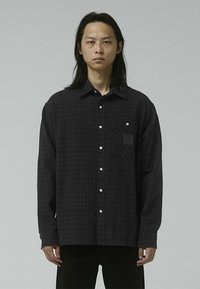 Black checkered shirt made from soft fabric, featuring a button-down collar, front pocket, and white buttons. Structured, relaxed fit.
