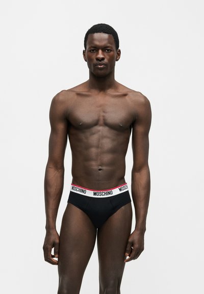 Μυώδης άνδρας με σκουρόχρωμο δέρμα, όρθιος, φοράει μαύρα εσώρουχα με λευκή ζώνη στη μέση με την ένδειξη «Moschino» σε απλό φωτεινό φόντο.