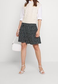 Gilet en tricot crème par-dessus une chemise blanche à col, associé à une jupe noire à pois blancs et des talons blancs à brides, tenant un petit sac blanc.