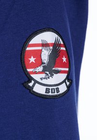 Blått tyg med ett broderat märke som visar en örn, röda ränder, två stjärnor och texten "BOB" nedanför. Cirkulärt mönster och texturerad detaljering.