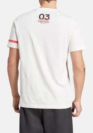 Mężczyzna w białej koszulce Adidas z napisem "03 Adidas Three Stripes Since 1949" na górnej części pleców oraz czerwoną opaską na lewym rękawie, w połączeniu z czarnymi szortami.