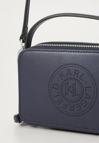 Geantă din piele bleumarin cu închidere cu fermoar, cu un design perforat al logo-ului pe față, curea subțire reglabilă și textură netedă.