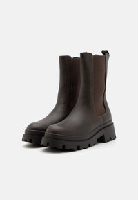 Bottines en cuir marron avec des panneaux élastiques sur les côtés et une semelle épaisse et texturée. Elles disposent d'une languette à l'arrière pour un enfilage facile.