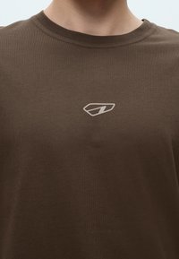 Κοντινό πλάνο ενός ατόμου που φοράει καφέ t-shirt με στρογγυλή λαιμόκοψη και μικρό φωτεινό γεωμετρικό λογότυπο στο κέντρο του στήθους.
