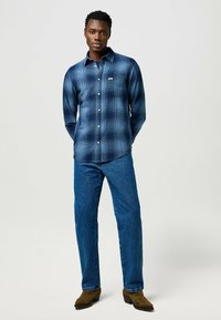 Camisa de cuadros azul de botones con mangas largas, bolsillo a la izquierda, usada con jeans azules y botas marrones. Textura de tela suave y diseño ajustado.