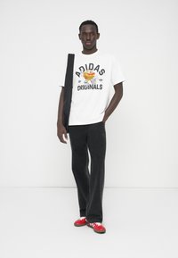 Balti marškinėliai su juodu užrašu „Adidas Originals“, tamsūs džinsai, raudoni batai su baltais akcentais, stovintis priešais neutralų foną.