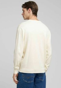 Maglione crema a maniche lunghe con scollo rotondo e polsini a coste, abbinato a jeans blu. Il tessuto appare morbido e leggero.