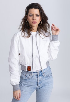 Veste bomber blanche courte avec des poignets côtelés gris, fermeture éclair et étiquette cousue à l'avant. Associée à un jean taille haute bleu clair.