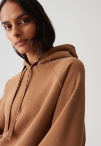 Close-up van een persoon met een gemiddelde huidtint en donker haar, gekleed in een bruin hoodie met trekkoorden, tegen een effen achtergrond.