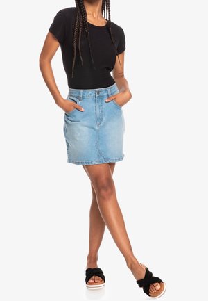 Femme portant un haut noir à manches courtes, jupe en jean bleu clair, mains dans les poches, sandales noires à enfiler, debout jambes croisées.