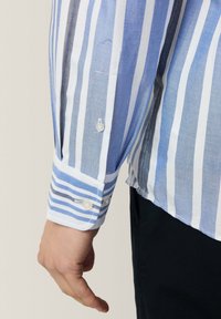 Hemd mit langen Ärmeln, vertikalen Blau- und Weißstreifen und einem weißen Manschette mit zwei Knöpfen. Der Stoff hat eine dezente Textur.