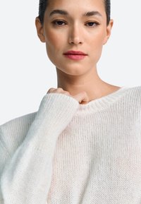 Lahka, kremno obarvana pletena majica z ohlapnim krojem, rebrastimi manšetami in širokim izrezom, ki poudarja teksturirano tkanino.